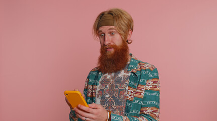 Happy excited joyful hippie man in pattern shirt use mobile cell phone typing browsing shouting say...