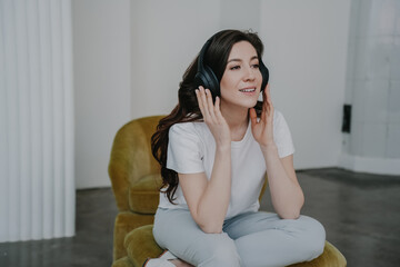 Beautiful female with loose wavy hair  in casual, relaxing at home listening music, using headphones, looking aside sits on chair. Attractive hispanic housewife smiling. Domestic leisure. Relaxation