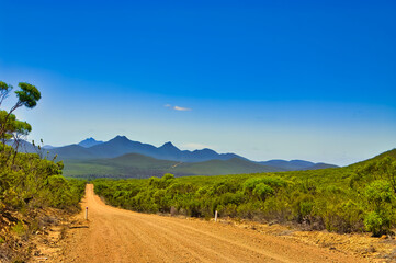 Fototapeta na wymiar Dirt road through the wilderness, leading to the sharply defined mountains of Stirling Range national park, Western Australia, on a sunny summer day 