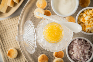 Gelber Saiblings Kaviar auf Eis in Glas Schale mit Porzellan Löffel, gekochte Eier Würfel, Charlotten Zwiebel Würfel, Toast Brot und Saure Sahne auf Silber Teller, Serviette und Marmor hell