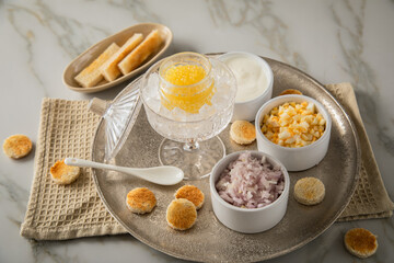 Gelber Saiblings Kaviar auf Eis in Glas Schale mit Porzellan Löffel, gekochte Eier Würfel, Charlotten Zwiebel Würfel, Toast Brot und Saure Sahne auf Silber Teller, Serviette und Marmor hell