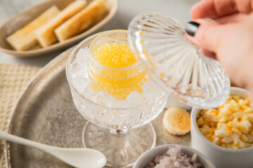 Gelber Saiblings Kaviar auf Eis in Glas Schale mit Porzellan Löffel, gekochte Eier Würfel, Charlotten Zwiebel Würfel, Toast Brot und Saure Sahne auf Silber Teller, Serviette und Marmor hell