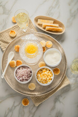 Gelber Saiblings Kaviar auf Eis in Glas Schale mit Porzellan Löffel, gekochte Eier Würfel, Charlotten Zwiebel Würfel, Toast Brot, Saure Sahne und Champagner Sekt auf Silber Teller und Marmor hell