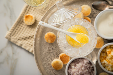 Gelber Saiblings Kaviar auf Eis in Glas Schale mit Porzellan Löffel, gekochte Eier Würfel, Charlotten Zwiebel Würfel, Toast Brot, Saure Sahne und Champagner Sekt auf Silber Teller und Marmor hell