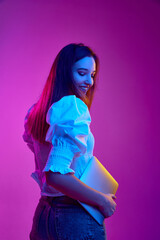 Portrait of young girl, student in white blouse posing with laptop over purple background in neon...