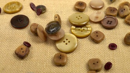 Brown buttons in different designs and sizes for sewing on clothes fall on brown burlap, slow motion