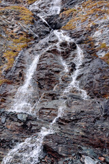 Small mountain waterfall flows over granite ledges of alpine rocks covered with yellowed autumn mosses and lichens, vertical shot, Cogne, Aosta Valley, Italy