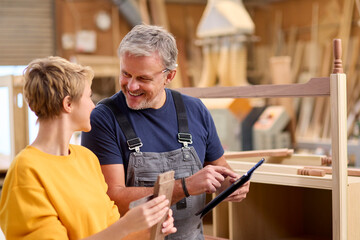 Female Apprentice Learning From Mature Male Carpenter With Digital Tablet In Furniture Workshop