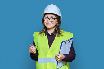 Professional woman in safety vest helmet holding clipboard on blue background
