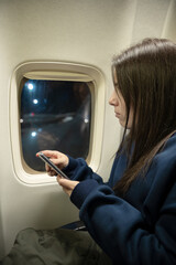 Young woman sitting in an airplane while looking out of the window and using her smartphone - travel concept - obrazy, fototapety, plakaty