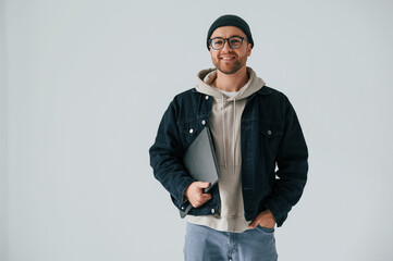 IT specialist in hat and glasses standing with laptop. Handsome man is in the studio against white...
