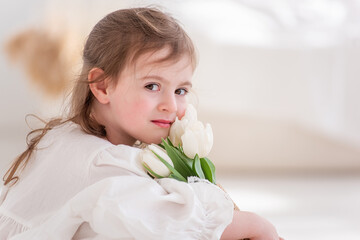 Close-up portrait of a little girl in a white dress, nightgown. Toddler embraces a bouquet of fresh