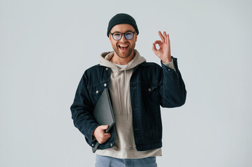 IT specialist in hat and glasses standing with laptop. Handsome man is in the studio against white background