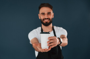 Giving the cup of coffee. Cafe worker is in apron. Handsome man is in the studio against blue background