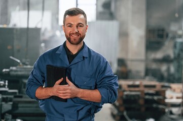 Smiling and holding tablet in hands. Factory worker in blue uniform is indoors