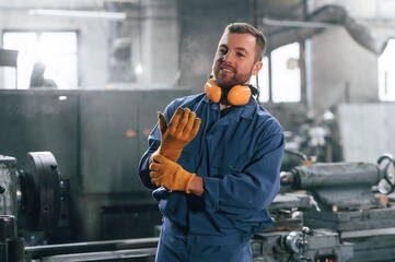 With gloves and protective headphones. Factory worker in blue uniform is indoors