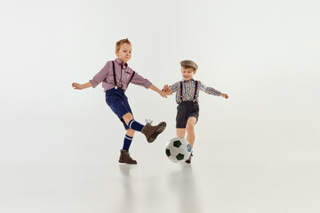 Fototapeta na wymiar Competition. Boys, children in classical retro clothes playing football over grey studio background. Concept of game, childhood, friendship