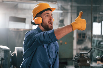 Man in hard hat and headphones is showing thumb up. Factory worker in blue uniform is indoors