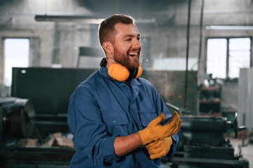 In yellow colored hear protective headphones and gloves. Factory worker in blue uniform is indoors