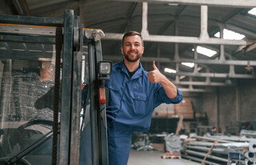 Fototapeta na wymiar In the forklift, showing thumb up. Factory worker in blue uniform is indoors