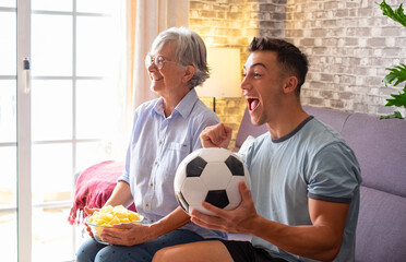 Joyful young boy and senior grandmother soccer fans watching a football game on tv sitting on a...
