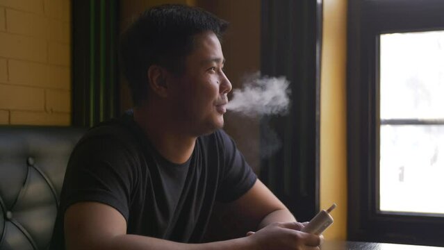Portrait of a man smoking a tobacco heating device at a table by the window in a cafe. Close-up of a cheerful guy sitting with an electronic cigarette in his hand.