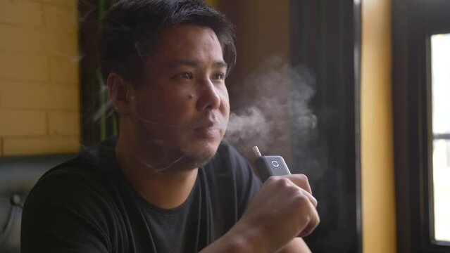 
Portrait of a man smoking a tobacco heating device at a table by the window in a cafe. Close-up of a sad guy sitting with an electronic cigarette in his hand.
