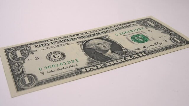 One new one dollar bill lies on a white surface. The camera rotates around the banknote.