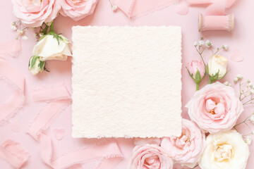 Blank square card between pink roses and pink silk ribbons on pink top view, wedding mockup
