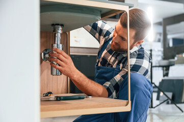 Busy by repairing siphon. Plumber in blue uniform is at work in the bathroom