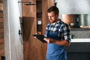 Standing and holding notepad. Plumber in blue uniform is at work in the bathroom