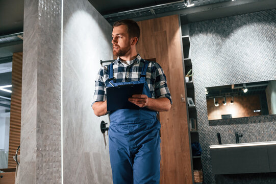 Standing and holding notepad. Plumber in blue uniform is at work in the bathroom