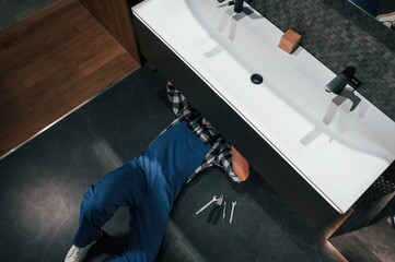 Lying down on the floor, fixing the siphon. Plumber in blue uniform is at work in the bathroom