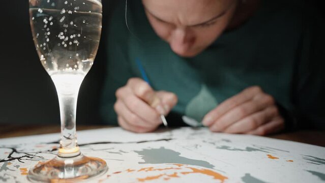 The girl paints the picture by numbers with paint and brushes, the camera focuses on a glass of champagne in the foreground.