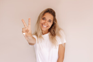 A young attractive caucasian smiling cheerful blonde woman in a white t-shirt shows a peace gesture with her hand isolated on a beige background. Girl shows a victory sign