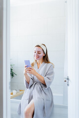 Smiling woman with facial pink clay mask on the face using smartphone at home bath enjoying relaxation and spa beauty treatment. Beauty blogger creating content. Vertical. Selective focus, copy space