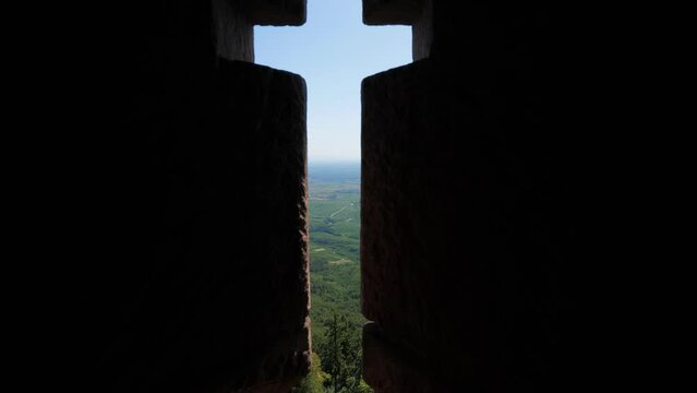 Camera move to narrow embrasure and look out, typical arbalestina (crosslet loop or firing slot) in stone wall of old castle. Fortification located on top of mountain, overlooking green plain