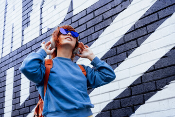 Stylish woman in blue jacket and heart shaped sunglasses wearing wireless headphones and listening to music while walking near city urban painted wall. Fashionable hipster lifestyle. Selective focus