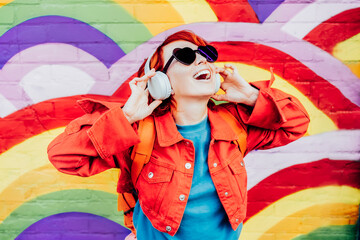Emotional stylish woman in bright clothes wearing wireless headphones, listening to music and singing on rainbow graffity on the brick wall background. Fashionable hipster lifestyle - obrazy, fototapety, plakaty