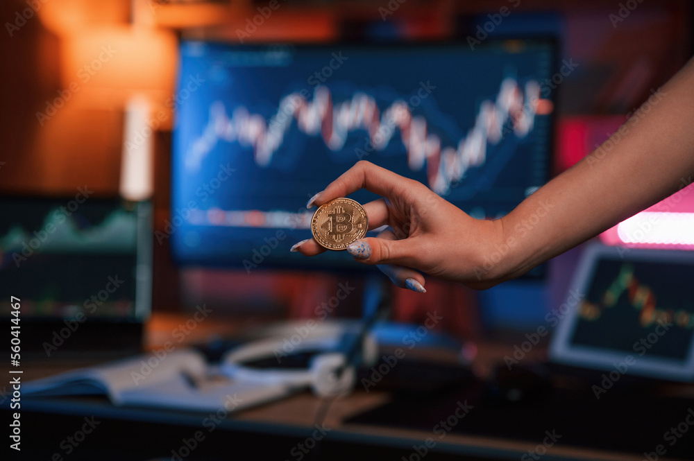 Poster close up view of woman's hand that holding bitcoin against computer display and tablet with graphs