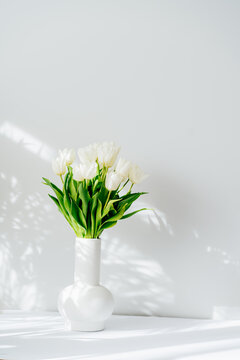 Spring bouquet of white tulip flowers in vase stand on white table near light grey wall with highlights and shadows. Gift for holiday, birthday, 8 March, Mother's Day. Vertical card. Selective focus