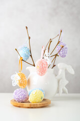 Home interior with easter decor. Vase with willow tree branches with Easter eggs and bunny on white table and background with copy space.
