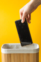 Man's hand putting smartphone into wooden basket on a yellow background. Dependance and digital detox concept.