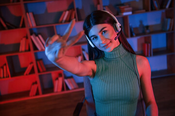 Positive young woman in headphones is sitting by a pc. Neon lighting
