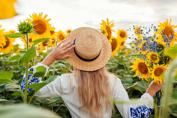 Young stylish woman walking in blooming sunflower field at sunset picking flowers. Ethnic clothing...