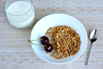 granola with milk in bowl decorated with cherry and glass of milk and teaspoon, close-up