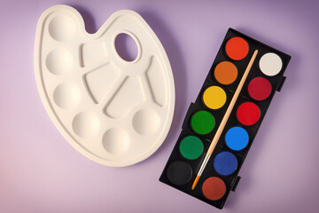 Brand new watercolor paints, brush and plastic palette on a purple background. Drawing, arts and...