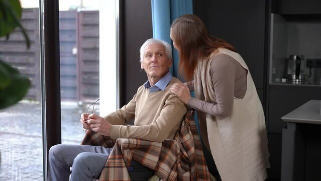 Senior Caucasian woman pushing wheelchair with man to window talking smiling. Loving caring wife enjoying leisure with husband at home indoors. Bonding and lifestyle