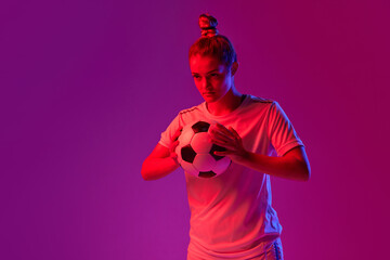 Concentrated young girl, professional female football, soccer player posing with ball over gradient...
