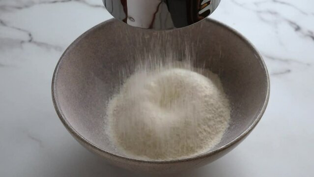 Wheat flour sifting through a metal sieve and pouring into ceramic mixing bowl. Shortcrust pastry preparation. Ingredients for shortbread dough. Close up. Marble background
